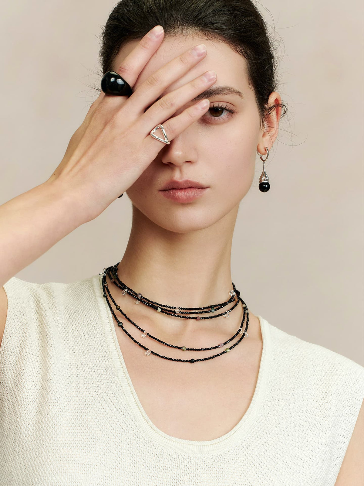 Model wearing Fairyokai Starlight Chain multi-layer necklace wrapped several times around her neck, right hand covering her right eye, black earrings, white sleeveless top, and a black ring on her right finger.