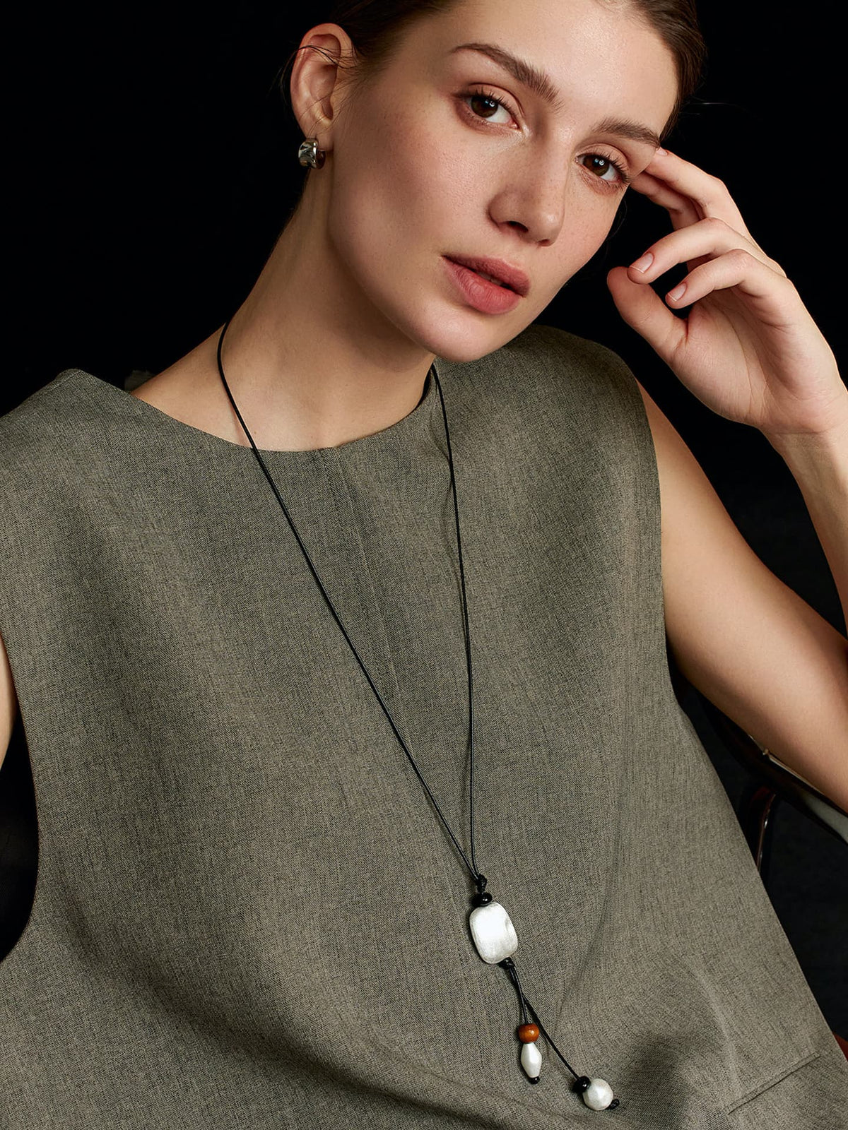 Model in a solid sleeveless top, leaning back on a chair, looking diagonally up toward the camera with one hand resting on her forehead, wearing the Fairyokai Suspended Wood & Waxed Cord Necklace, on black background.