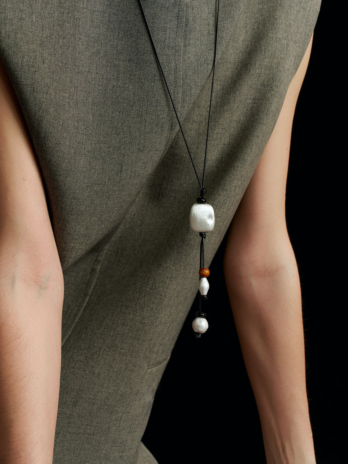 Model in a solid sleeveless top, wearing the Suspended Wood & Waxed Cord Necklace, with both hands resting on a table, on black background.