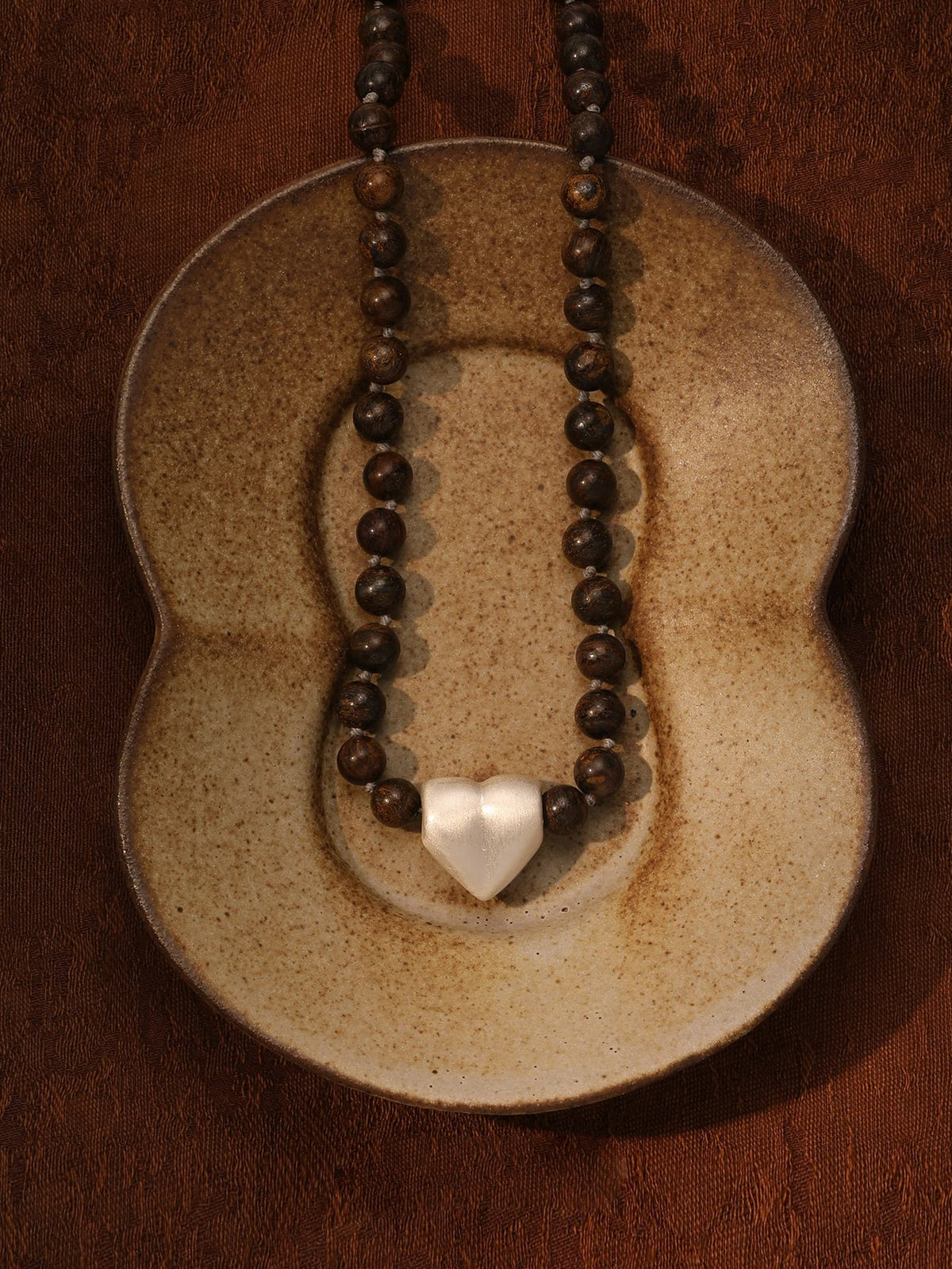 Fairyokai Tiger Eye and Silver Heart necklace laid flat on a brown figure-8 ceramic plate, top-down view.