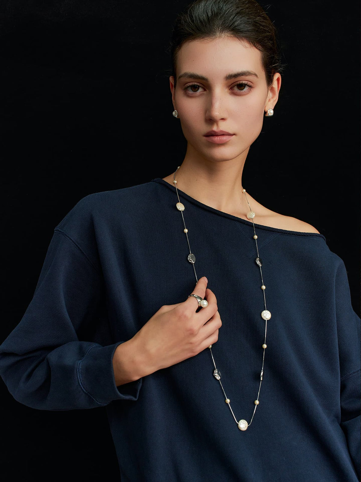 Model in a navy top, wearing the Fairyokai Tracing Light irregular faux pearl lobster clasp necklace, one hand touching the necklace, facing the camera on a black background.