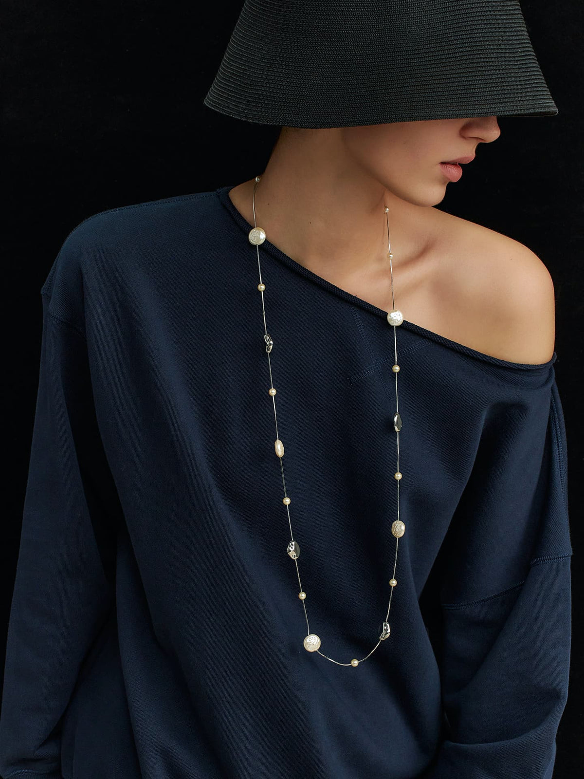 Model wearing a black hat covering her eyes, navy top with left shoulder exposed, looking to the left, wearing the Fairyokai Tracing Light irregular faux pearl lobster clasp necklace on a black background.