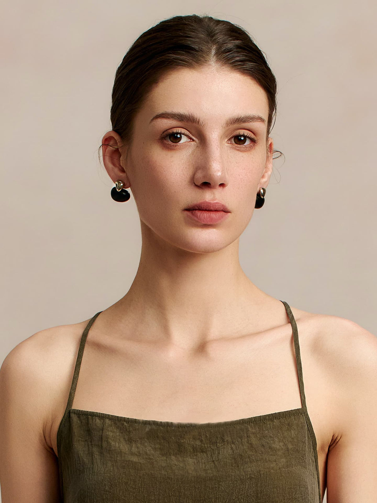 Model in a deep green thin-strap dress wearing Fairyokai Tranquil Ebony stream stone earrings, facing the camera — front view, against a beige background