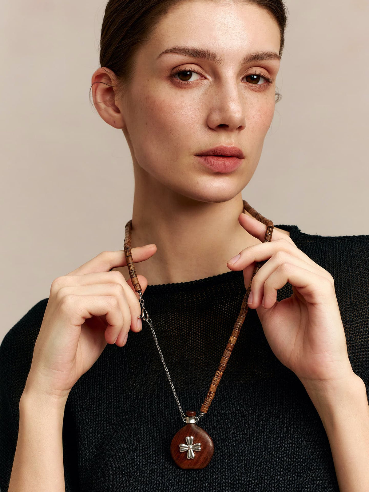Model in a black top, holding the Fairyokai Wooden Serenity wenge wood vase necklace with both hands slightly raised, facing the camera, on neutral background.