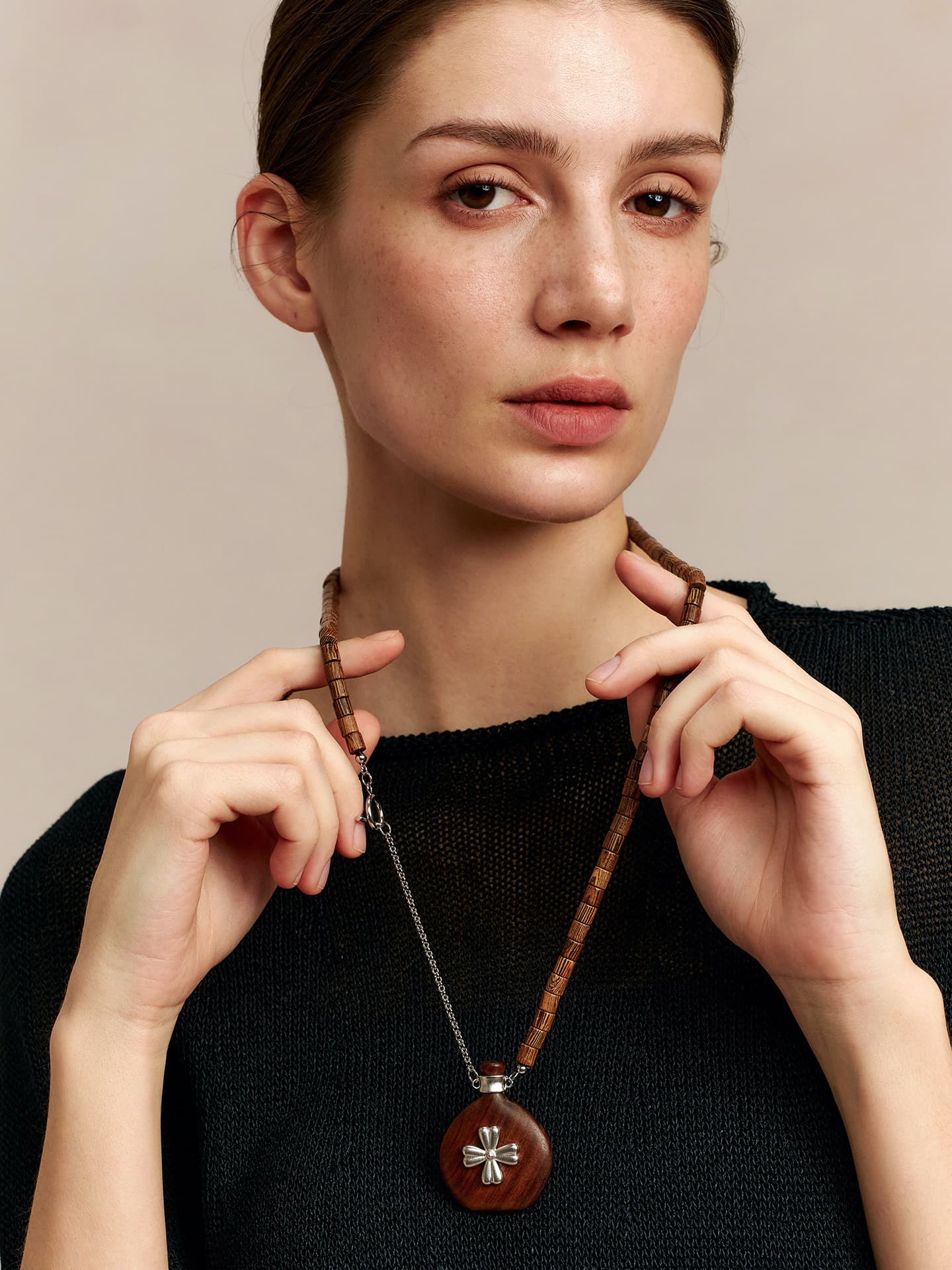 Model in a black top, holding the Fairyokai Wooden Serenity wenge wood vase necklace with both hands slightly raised, facing the camera, on neutral background.