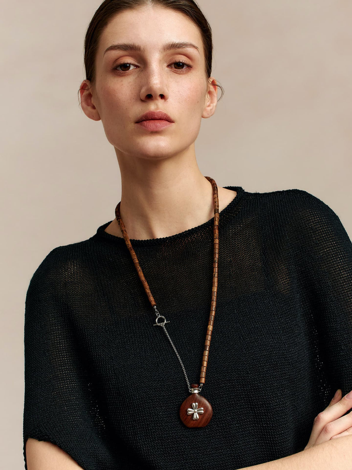 Model in a black top, facing the camera with arms crossed over her chest, wearing the Fairyokai Wooden Serenity wenge wood vase necklace, on neutral background.